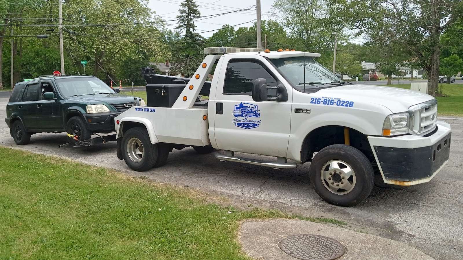 ALDM Management's white wrecker, blue shield logo on the door and the phone number on the hood, with a green SUV hitched on the back, parked at a Cleveland curb.