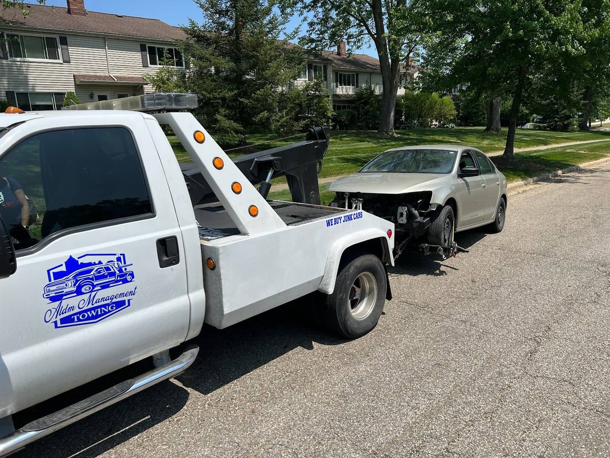 ALDM Management's white wrecker with the blue shield logo on the door, a silver sedan with front-end damage hitched on the back, on a residential street with houses and trees.