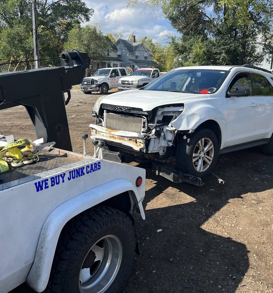 A white SUV with a missing front bumper and exposed radiator, hitched to the back of a tow truck whose fender reads WE BUY JUNK CARS, in a dirt lot.