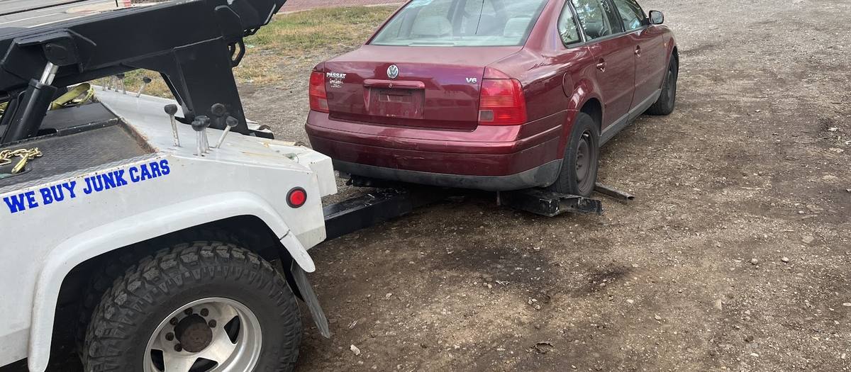 A maroon Volkswagen sedan with its front wheels strapped to the tow rig of a white truck lettered WE BUY JUNK CARS, in a gravel lot with a street beyond.
